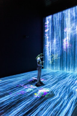 A person stands in a dark room surrounded by digital projections of cascading blue vertical lines resembling a waterfall, with colorful floral patterns scattered throughout. The room is illuminated by the vivid projections, creating an immersive and serene environment.