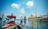 A man and a woman walk on a dock next to a body of water. A wooden boat with the number 15 and a Malaysian flag is docked nearby. In the background, there is an ornate mosque with a pink dome and tall minaret against a blue sky.