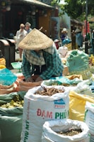 A person wearing a conical hat is seen working at an outdoor market surrounded by large sacks filled with various herbs and spices. The setting is vibrant and lively, with several people in the background, likely shoppers or other vendors. The area appears to be bustling with activity on a sunny day.