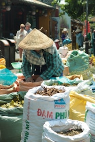 A person wearing a conical hat is seen working at an outdoor market surrounded by large sacks filled with various herbs and spices. The setting is vibrant and lively, with several people in the background, likely shoppers or other vendors. The area appears to be bustling with activity on a sunny day.