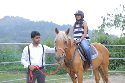 A woman wearing a helmet sits atop a brown horse in an outdoor setting. A man in a white shirt with suspenders stands beside them, holding the horse's reins. The background features greenery and distant hills under a bright sky.