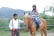 A woman wearing a helmet sits atop a brown horse in an outdoor setting. A man in a white shirt with suspenders stands beside them, holding the horse's reins. The background features greenery and distant hills under a bright sky.