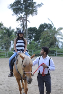A woman wearing a striped sleeveless top and jeans is sitting on a brown horse. She is wearing a protective helmet. A man in a white shirt with suspenders is walking beside the horse, holding a red lead rope. They are in an outdoor setting with a sandy ground and surrounded by tall trees and greenery.
