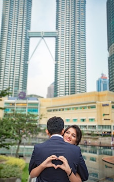 A couple stands closely together with the woman smiling at the camera, while the man, seen from the back, forms a heart shape with his hands. They are in an urban setting with tall skyscrapers in the background, and there is a reflection pool and some greenery nearby.