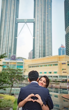 A couple stands closely together with the woman smiling at the camera, while the man, seen from the back, forms a heart shape with his hands. They are in an urban setting with tall skyscrapers in the background, and there is a reflection pool and some greenery nearby.