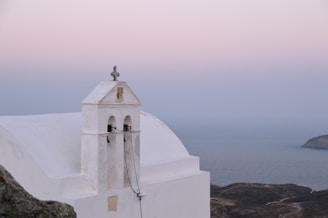 A serene landscape with a whitewashed building featuring a bell tower and a cross on top, set against a backdrop of soft pink and blue sky. The building is positioned on a hillside overlooking the calm sea, with rugged terrain visible in the distance.