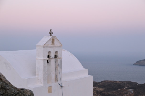 A serene landscape with a whitewashed building featuring a bell tower and a cross on top, set against a backdrop of soft pink and blue sky. The building is positioned on a hillside overlooking the calm sea, with rugged terrain visible in the distance.
