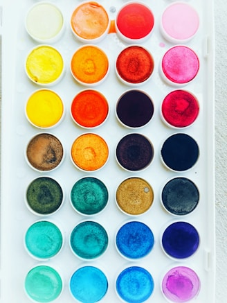 A neatly arranged set of watercolor paints in a white plastic tray, featuring multiple rows of circular wells with vibrant colors including shades of yellow, orange, red, pink, brown, blue, and green.