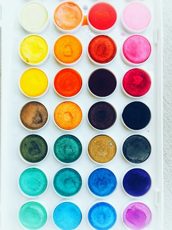 A neatly arranged set of watercolor paints in a white plastic tray, featuring multiple rows of circular wells with vibrant colors including shades of yellow, orange, red, pink, brown, blue, and green.