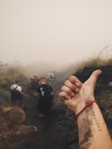 A group of people hiking on a misty trail with blurred visibility due to the fog. In the foreground, a close-up of a person's dirty hand and wrist is visible, featuring a tattoo and a red bracelet. The scene conveys a sense of adventure and unity.
