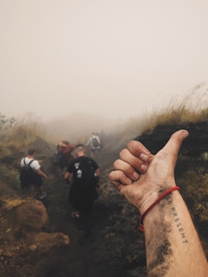 A group of people hiking on a misty trail with blurred visibility due to the fog. In the foreground, a close-up of a person's dirty hand and wrist is visible, featuring a tattoo and a red bracelet. The scene conveys a sense of adventure and unity.
