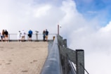 A group of people standing on a viewing platform overlooking a bright, cloudy sky. The photo focuses on a silver metal railing that leads towards the blurred figures in the background. The floor of the platform is made of light-colored stone.