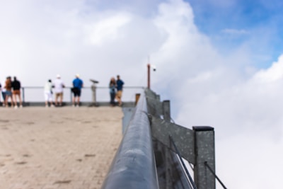 A group of people standing on a viewing platform overlooking a bright, cloudy sky. The photo focuses on a silver metal railing that leads towards the blurred figures in the background. The floor of the platform is made of light-colored stone.