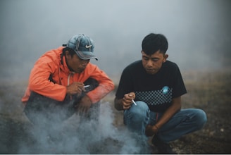 Two men are crouched on the ground in a misty or smoky environment. The man on the left wears an orange jacket and sports cap, while the man on the right is in a black t-shirt with a checkered pattern. Both are holding cigarettes and appear to be engaged in conversation or contemplation.