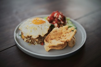A plate containing a serving of fried rice topped with a sunny-side-up egg, accompanied by slices of crispy fried chicken and a side of vegetables with sauce.
