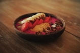 A vibrant smoothie bowl is presented on a wooden surface, featuring colorful toppings such as sliced bananas, watermelon chunks, apple slices, granola, seeds, and a dollop of nut butter.