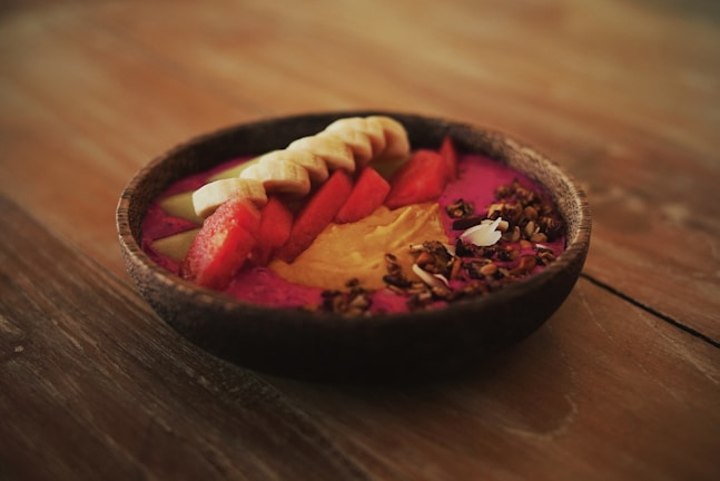 A vibrant smoothie bowl is presented on a wooden surface, featuring colorful toppings such as sliced bananas, watermelon chunks, apple slices, granola, seeds, and a dollop of nut butter.
