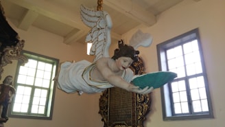 A detailed sculpture of an angel with outstretched arms holding a green cushion is suspended from the ceiling. The angel has white wings, and its attire is white with gold accents. The setting appears to be inside a building with large windows and ornate architectural details.