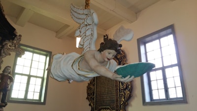 A detailed sculpture of an angel with outstretched arms holding a green cushion is suspended from the ceiling. The angel has white wings, and its attire is white with gold accents. The setting appears to be inside a building with large windows and ornate architectural details.