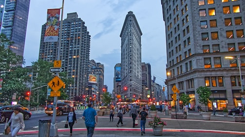 In an urban setting, a bustling street leads up to a prominent triangular-shaped skyscraper, with people walking and vehicles visible. Surrounding the main building, various high-rise buildings exhibit a mix of architectural styles. Street signs and traffic lights are evident, and the scene conveys the bustling nature of city life.