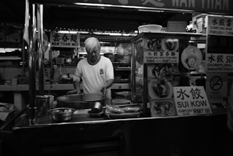A street food vendor is preparing food at a stall with various signs displaying menu items like dumplings. The vendor has white hair and is wearing a white shirt. In the foreground, there are cooking utensils and containers, while in the background, the stall interior is visible with cooking equipment and a fan.