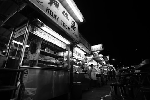A bustling street food market at night featuring a row of illuminated food stalls. Signs with various names and menus can be seen above the stalls. People are gathered around, some standing near the counters while others walk through the marketplace.