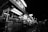 A bustling street food market at night featuring a row of illuminated food stalls. Signs with various names and menus can be seen above the stalls. People are gathered around, some standing near the counters while others walk through the marketplace.
