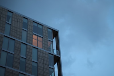 A modern building with a facade composed of large glass windows and vertical panels. One window is illuminated with a warm light, contrasting against the cool tones of the dusk sky.