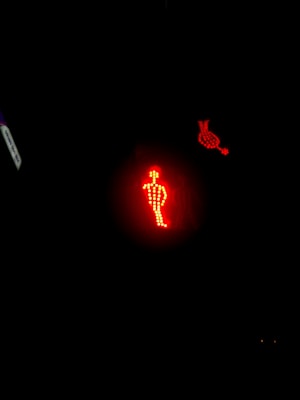 A red LED pedestrian traffic light is illuminated in a dark setting. The traffic light displays the figure of a standing person made from small red dots, indicating not to cross. Nearby, there is another illuminated symbol made of the same red LED dots.