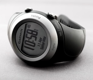 A sleek and modern digital wristwatch displaying the time 10:58. The watch has a round face with a metallic silver and black casing. Various buttons are visible on the sides, labeled 'training', 'lap/reset', and 'menu'. The watch is on a light gray, textured background.