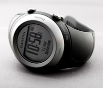 A sleek and modern digital wristwatch displaying the time 10:58. The watch has a round face with a metallic silver and black casing. Various buttons are visible on the sides, labeled 'training', 'lap/reset', and 'menu'. The watch is on a light gray, textured background.