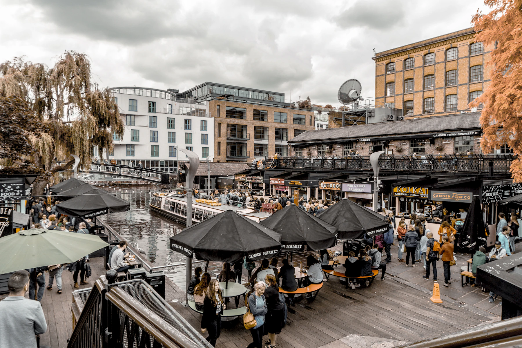 Photograph of People sit beside the canal among the brick buildings, signs and market terraces of Camden Lock.