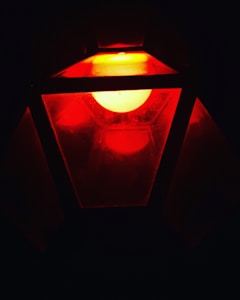 A red light source is enclosed within a geometric, lantern-like structure, casting an intense glow. The light creates a warm, mysterious atmosphere and the angular panels suggest a modern design.