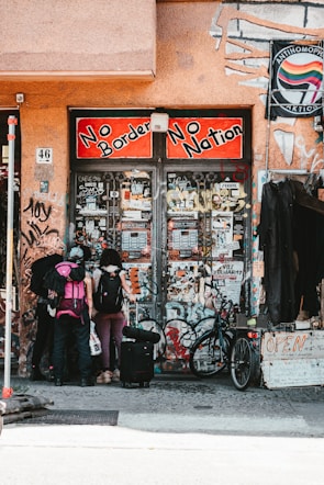 A group of three people with backpacks and luggage stand in front of a graffiti-covered building entrance. The sign above reads 'No Borders No Nation' in bold red and black lettering. Various posters and stickers cover the walls and doors. A bicycle is parked nearby, and a sign with 'OPEN' is visible on the right.