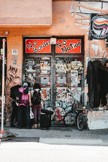 A group of three people with backpacks and luggage stand in front of a graffiti-covered building entrance. The sign above reads 'No Borders No Nation' in bold red and black lettering. Various posters and stickers cover the walls and doors. A bicycle is parked nearby, and a sign with 'OPEN' is visible on the right.
