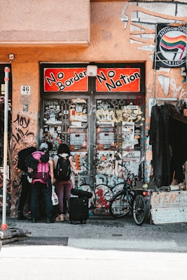 A group of three people with backpacks and luggage stand in front of a graffiti-covered building entrance. The sign above reads 'No Borders No Nation' in bold red and black lettering. Various posters and stickers cover the walls and doors. A bicycle is parked nearby, and a sign with 'OPEN' is visible on the right.