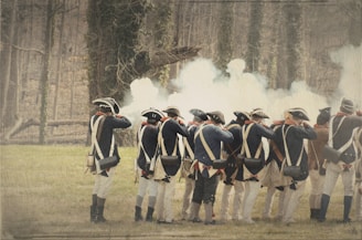 A group of historical reenactors in period military uniforms, standing in formation and firing muskets. Dense smoke clouds rise from the gunfire, partially obscuring the forested background. The soldiers wear blue coats, tricorn hats, and carry cartridge boxes.