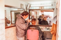 A barber is cutting a customer's hair in a small, vintage barber shop. The customer sits on a red barber chair, while the barber concentrates on the haircut. Both of them are visible in the large mirror on the wall. The shop is decorated with patterned tiles and has a few hanging items like a towel and a jacket. The lighting is warm and natural.