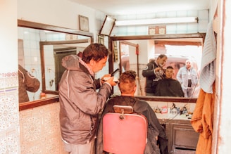 A barber is cutting a customer's hair in a small, vintage barber shop. The customer sits on a red barber chair, while the barber concentrates on the haircut. Both of them are visible in the large mirror on the wall. The shop is decorated with patterned tiles and has a few hanging items like a towel and a jacket. The lighting is warm and natural.