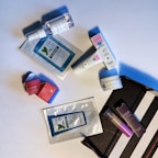 A collection of cosmetic and skincare products laid out on a white surface. These include facial masks, creams, and serums from various brands. The items are casually arranged, with some still in their packaging. The scene includes both rectangular and cylindrical packages with colorful labels.