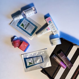A collection of cosmetic and skincare products laid out on a white surface. These include facial masks, creams, and serums from various brands. The items are casually arranged, with some still in their packaging. The scene includes both rectangular and cylindrical packages with colorful labels.