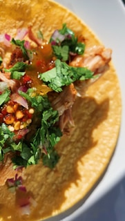 A close-up of a taco filled with shredded meat, topped with chopped cilantro, diced red onions, and a drizzle of sauce. The taco is on a corn tortilla, and the vibrant colors are enhanced by the lighting.