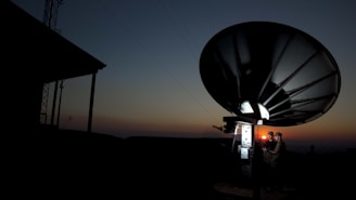 A large satellite dish is silhouetted against a dusk or dawn sky. Two individuals are standing near the base of the dish, illuminated by a small light, possibly engaged in some technical work or communication.
