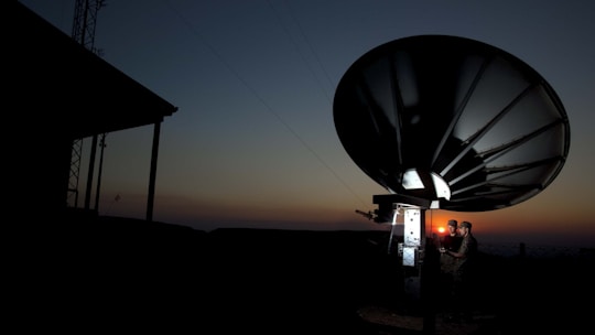 A large satellite dish is silhouetted against a dusk or dawn sky. Two individuals are standing near the base of the dish, illuminated by a small light, possibly engaged in some technical work or communication.