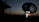 A large satellite dish is silhouetted against a dusk or dawn sky. Two individuals are standing near the base of the dish, illuminated by a small light, possibly engaged in some technical work or communication.