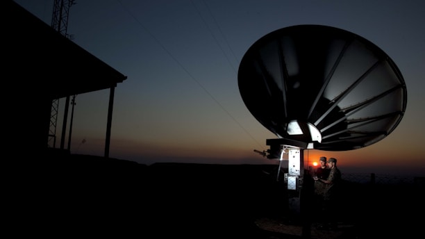 A large satellite dish is silhouetted against a dusk or dawn sky. Two individuals are standing near the base of the dish, illuminated by a small light, possibly engaged in some technical work or communication.