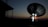 A large satellite dish is silhouetted against a dusk or dawn sky. Two individuals are standing near the base of the dish, illuminated by a small light, possibly engaged in some technical work or communication.