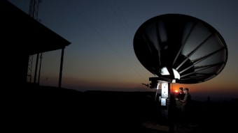 A large satellite dish is silhouetted against a dusk or dawn sky. Two individuals are standing near the base of the dish, illuminated by a small light, possibly engaged in some technical work or communication.