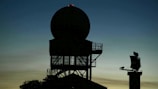 Silhouette of a radar tower with a large circular antenna against a dusk sky. The structure appears complex with metal framework and platforms. In the background, another smaller radar or communication device is visible on the right.