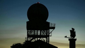 Silhouette of a radar tower with a large circular antenna against a dusk sky. The structure appears complex with metal framework and platforms. In the background, another smaller radar or communication device is visible on the right.